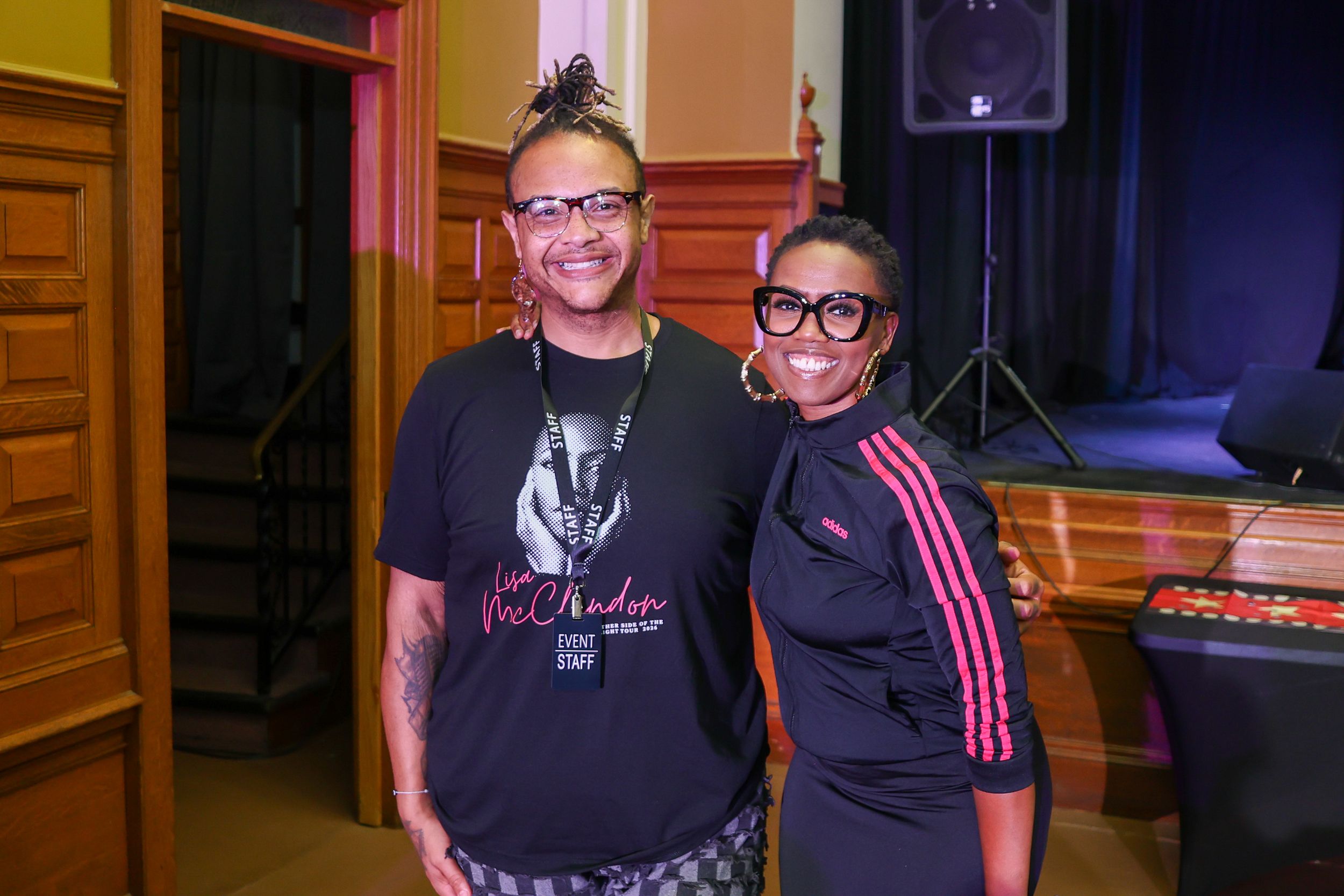 Two people pose together smiling at an indoor event, one wearing a graphic tee and the other a striped jacket.