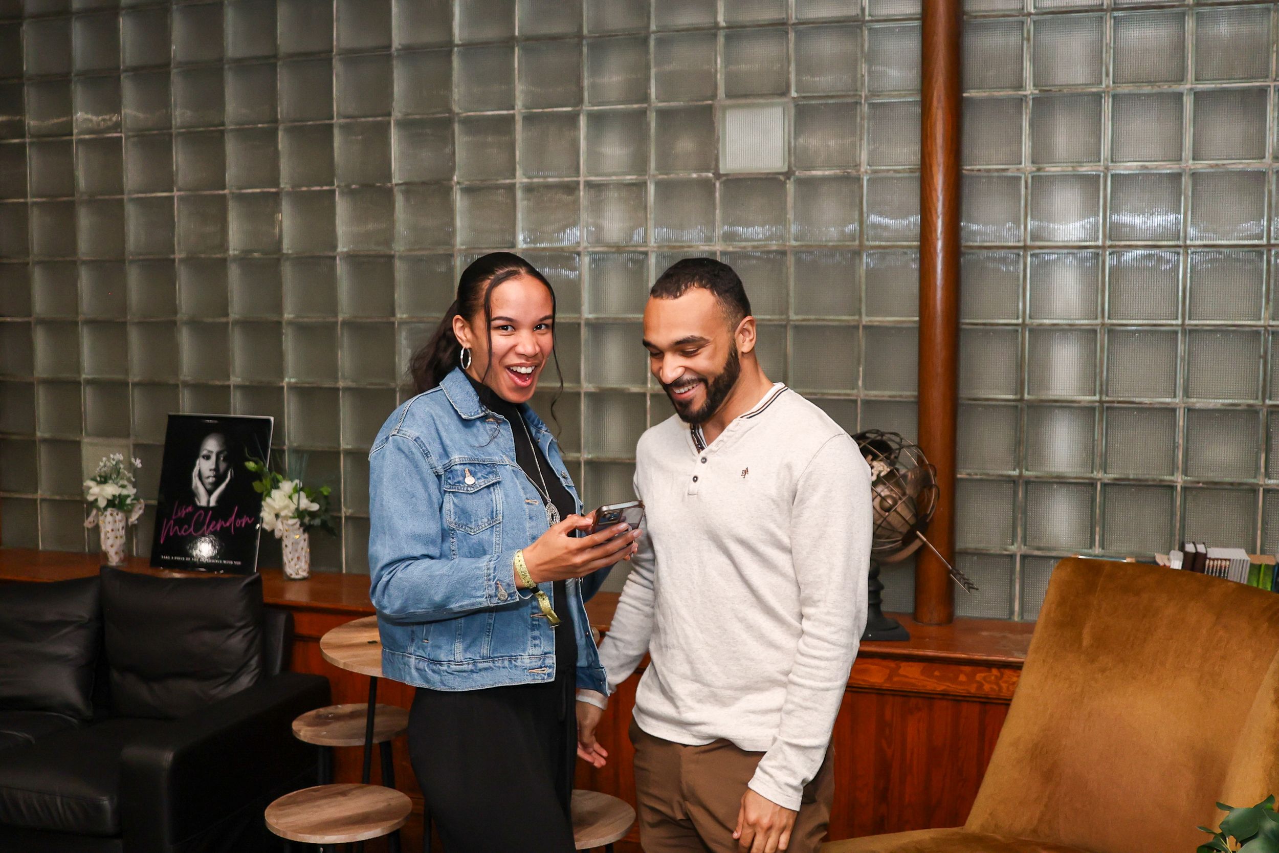 Two people smiling and looking at a smartphone together in a modern lounge with glass block walls.