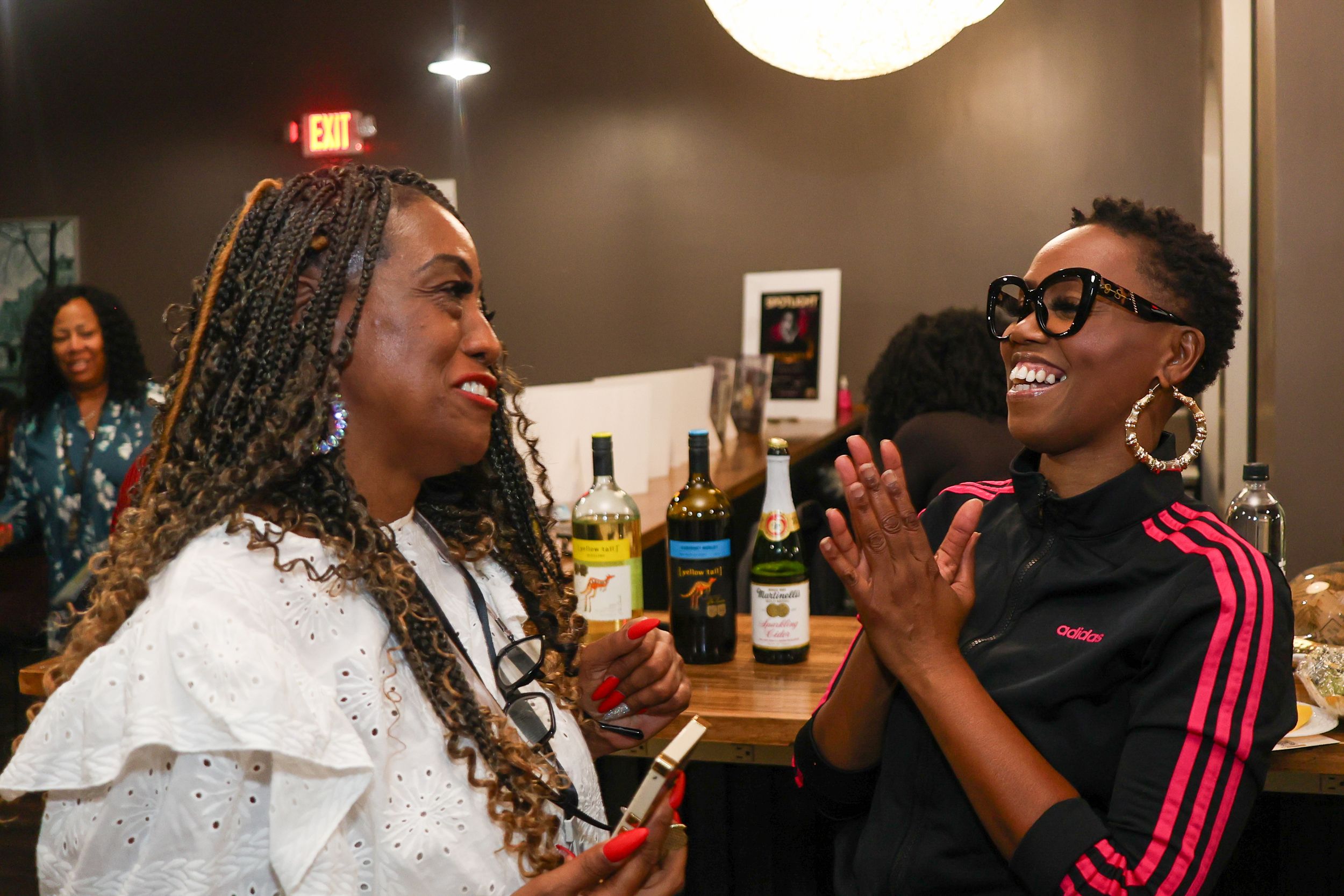 Two women laughing and socializing at an indoor gathering with wine bottles in the background.
