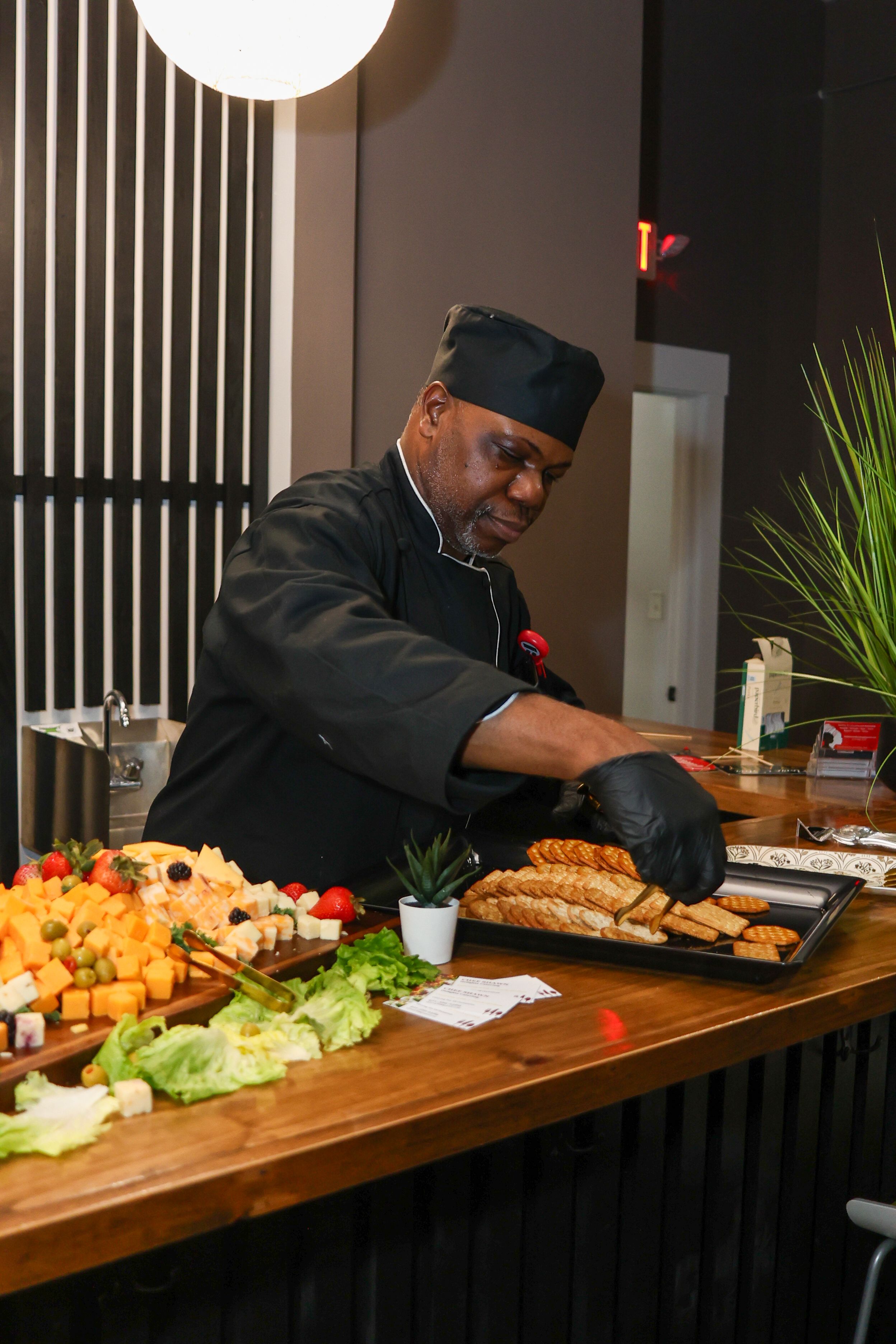 A chef in black uniform arranges food on a wooden buffet table with cheese, vegetables, and bread.
