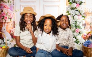 Three children with curly hair wearing hats and white shirts sitting together among floral decorations.