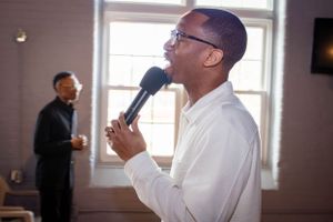 Speaker in a white shirt holds a microphone while performing in a bright room with windows.