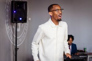 Speaker in glasses and white outfit gestures while speaking into microphone near sound equipment.