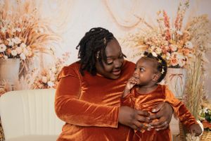 Two people in orange velvet outfits smile and embrace in front of floral backdrop.