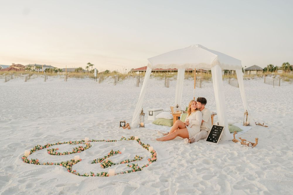 Romantic beach setup with white canopy tent and decorative candles arranged in heart shape on sand.