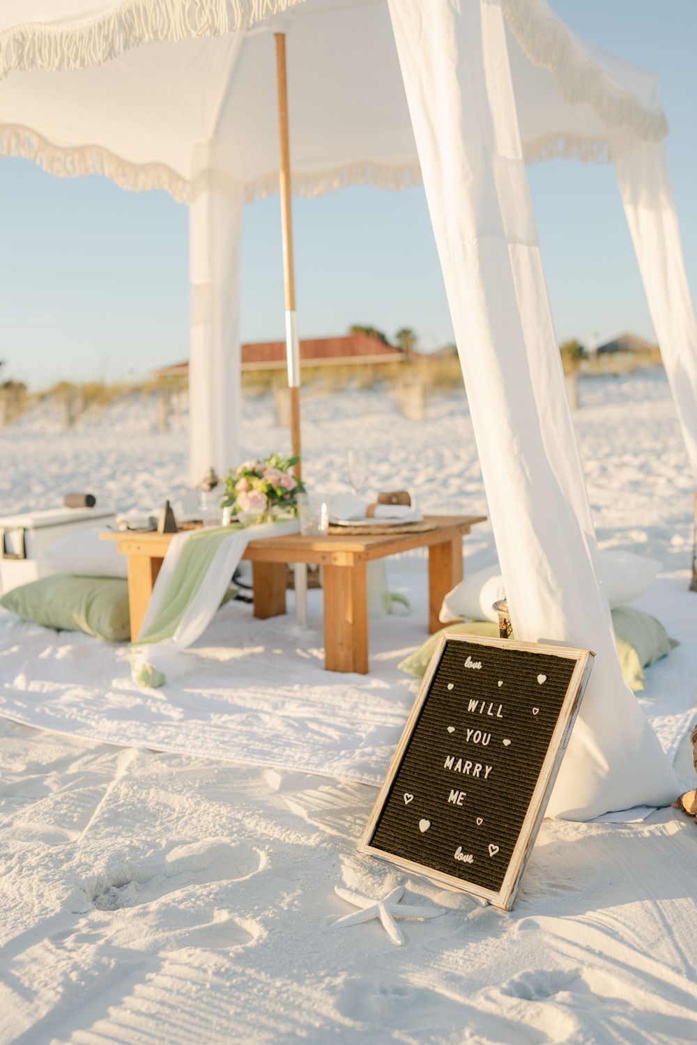 A romantic beach picnic setup with white canopy, wooden bench, and decorative details.