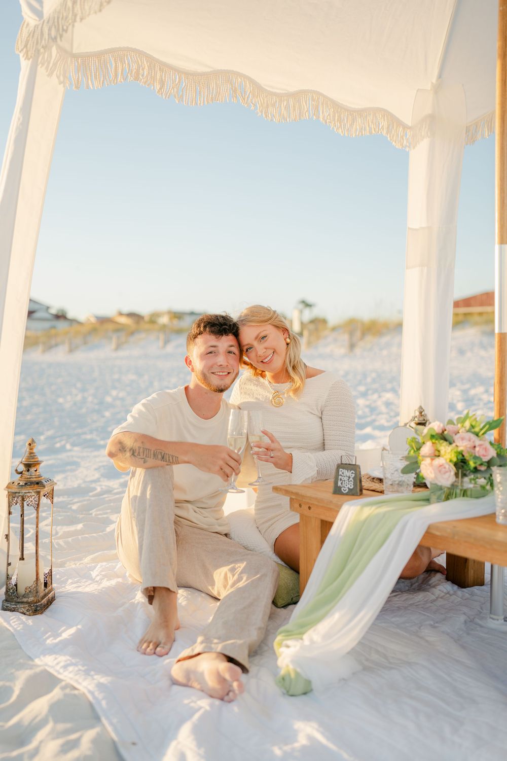 A couple sharing champagne under an elegant white beach canopy with floral decorations.