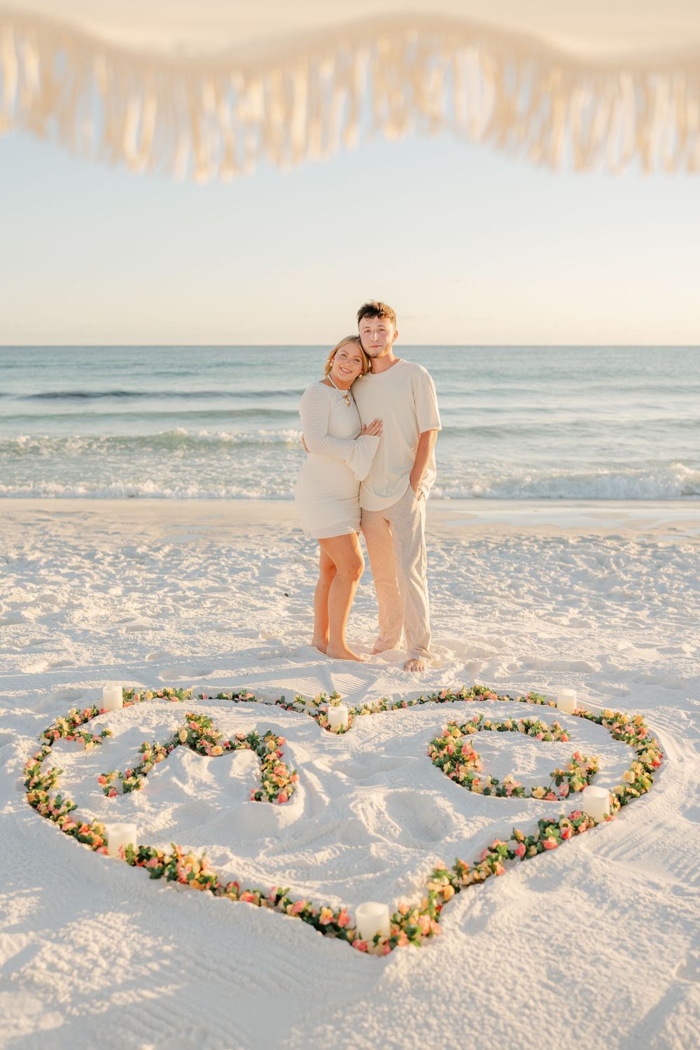 A romantic beach engagement photo with a floral heart design drawn in the sand.