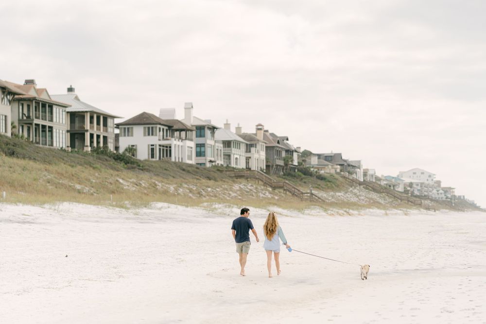 Couple walks with dog along beach with coastal homes on hillside in background.