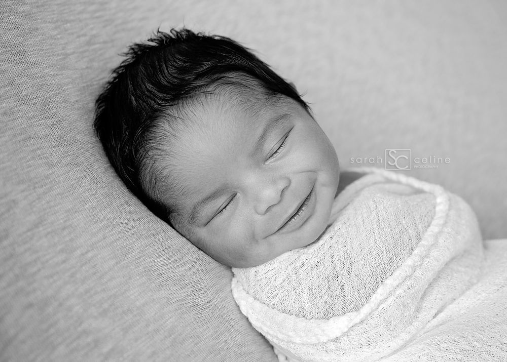 Peaceful sleeping newborn baby wrapped in white blanket smiling contentedly in black and white portrait.