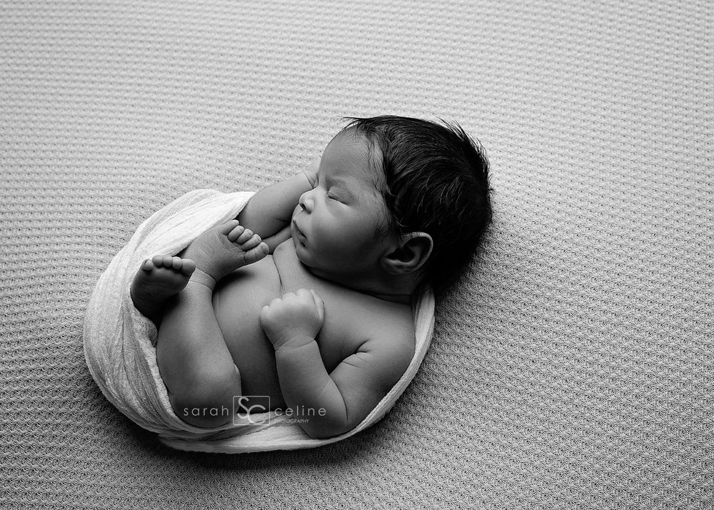 Black and white photograph of a newborn baby lying on a textured blanket in a curled-up sleeping position.