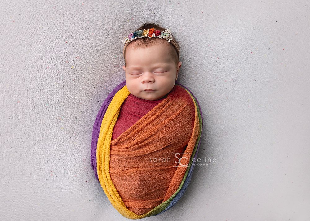 Sleeping newborn wrapped in autumn-colored swaddle blankets with floral headband on grey background.