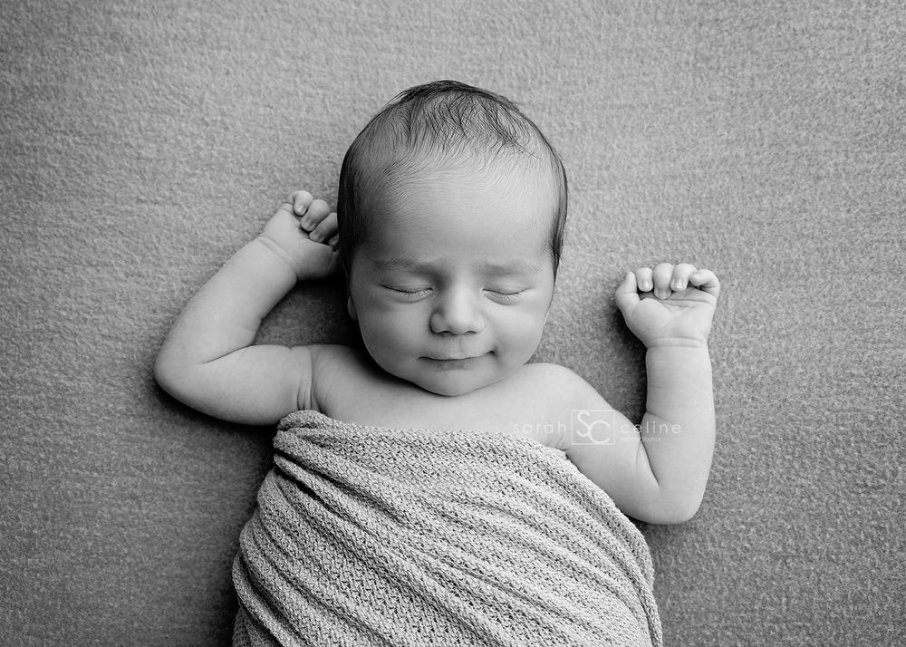 Sleeping baby wrapped in soft gray fabric with tiny fists raised lies peacefully on textured surface in black and white photo.