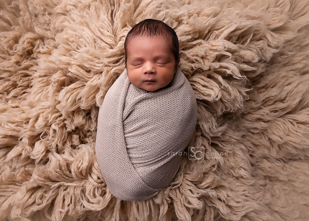 A newborn baby swaddled in a soft gray wrap rests peacefully on a fluffy beige fur blanket.