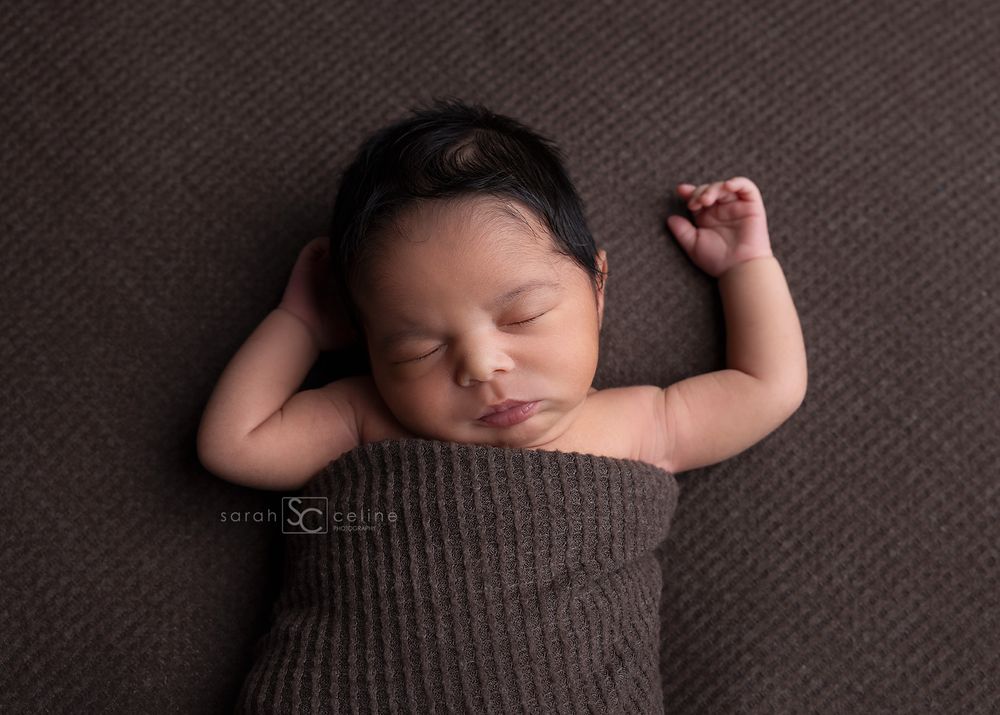 A peaceful sleeping newborn wrapped in a cozy brown knit blanket lies on a textured brown backdrop.
