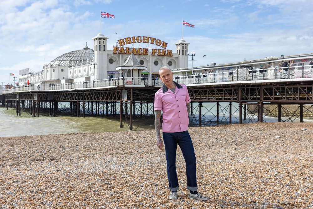 A man standing on a beach in front of the Brighton Palace Pier, wearing a pink shirt and blue pants.