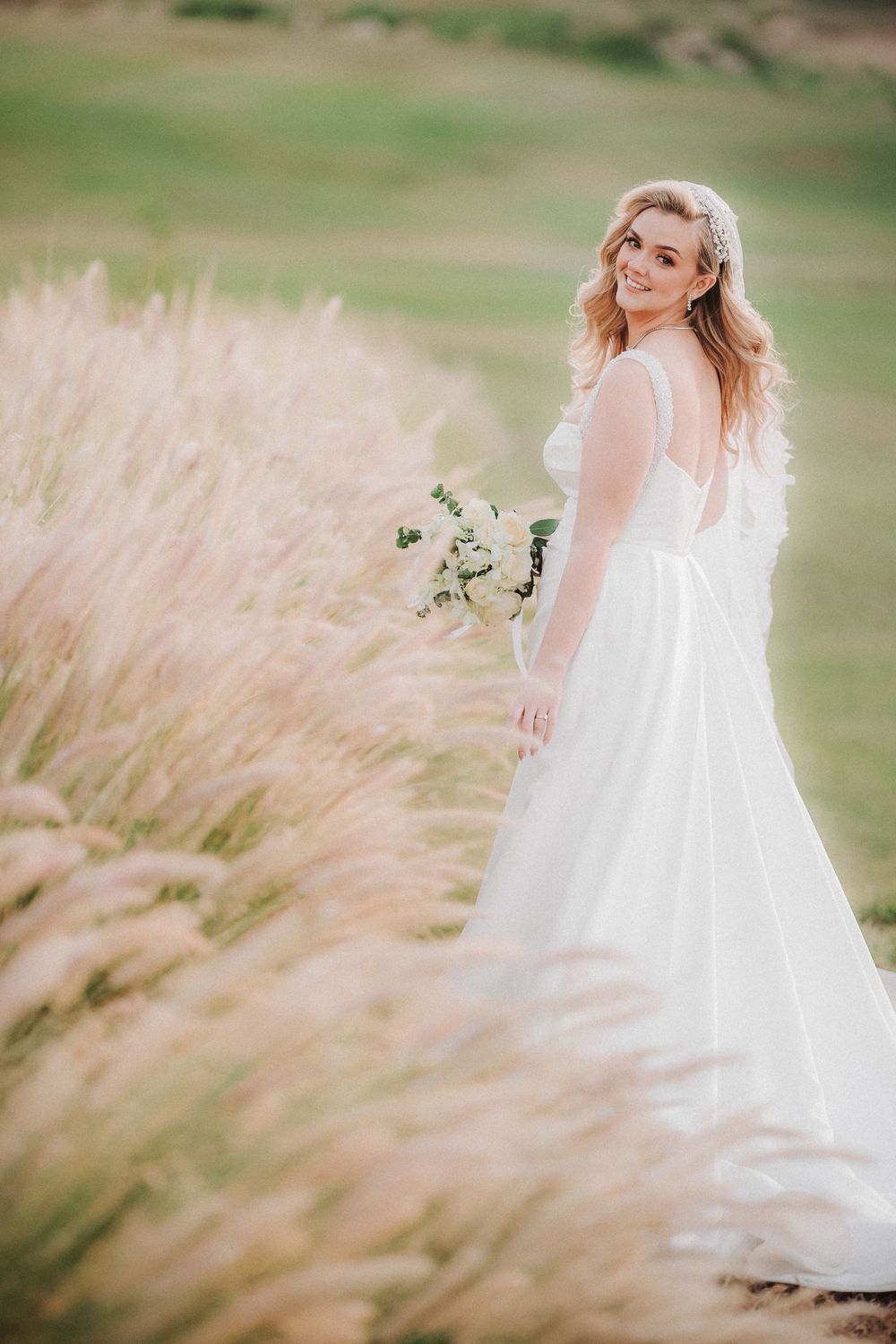 A bride in a flowing white dress poses in a field of pampas grass during golden hour.