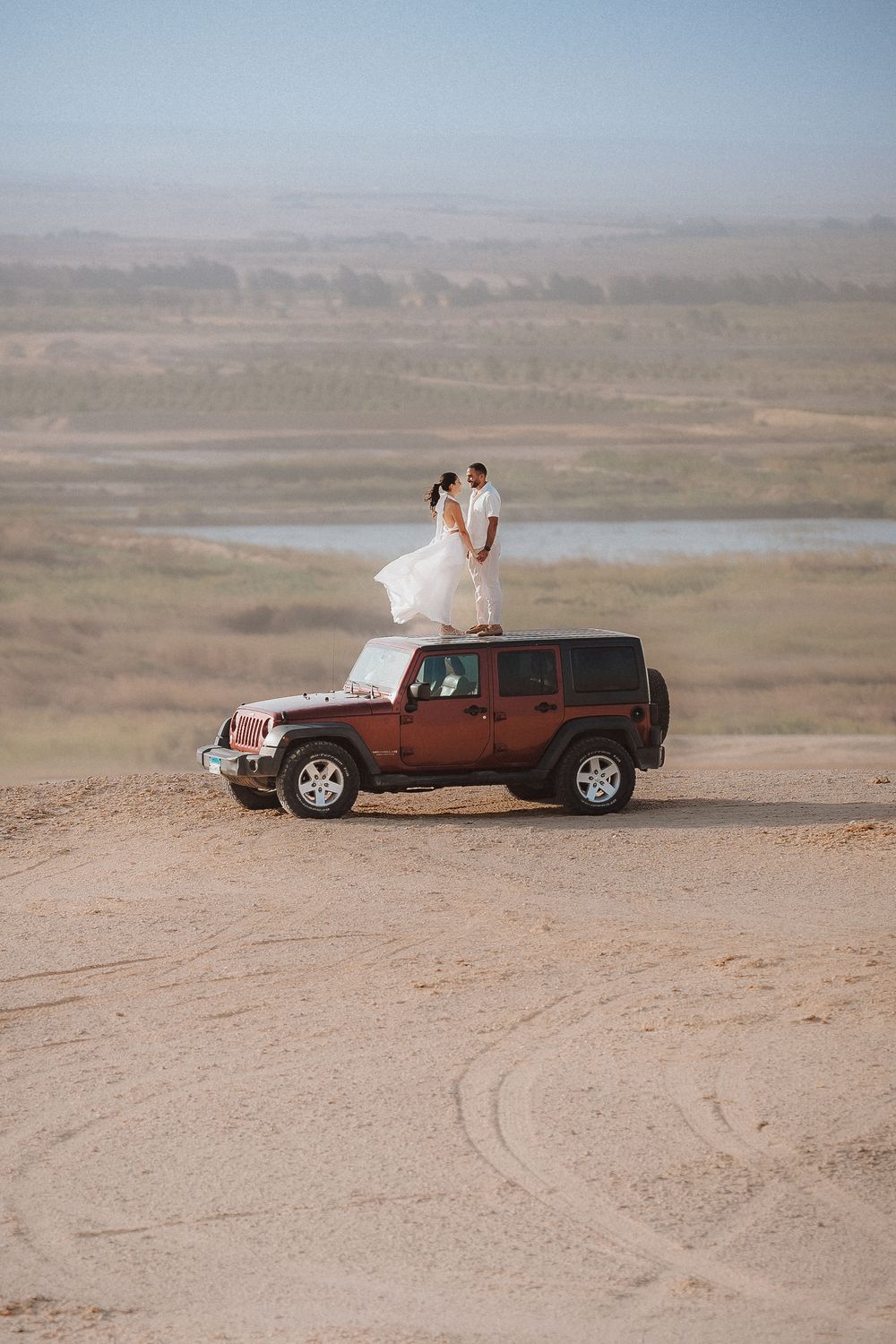 Wedding couple poses with vintage Jeep on desert cliff overlooking dramatic landscape.