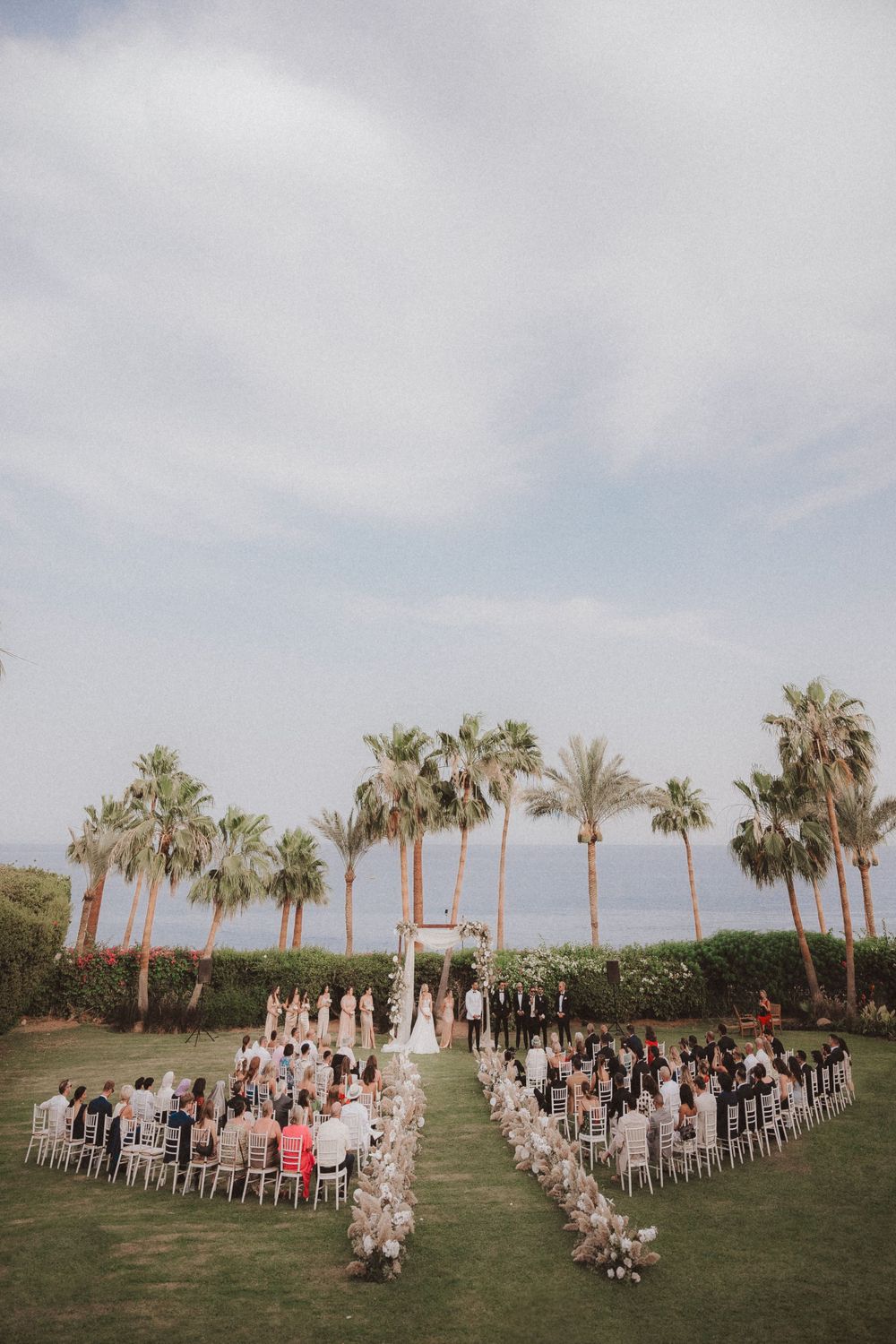 An outdoor wedding ceremony setup with palm trees and white chairs arranged in rows on a grassy lawn.
