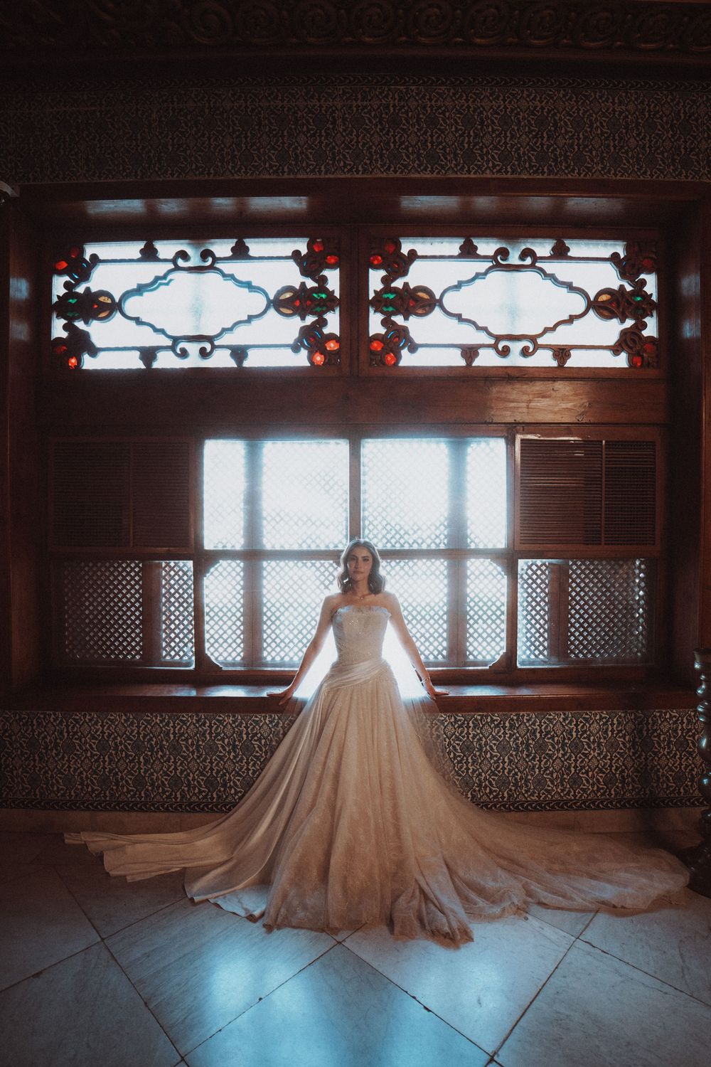 A flowing champagne-colored wedding gown with long train stands elegantly against ornate windows in a grand historic interior.