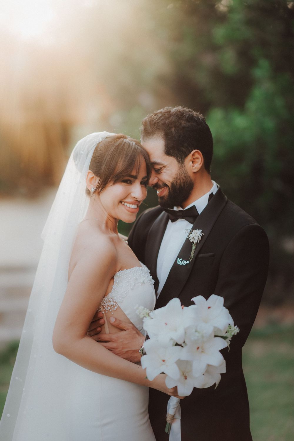 A romantic wedding couple embraces outdoors while holding a bouquet of white lilies at sunset.