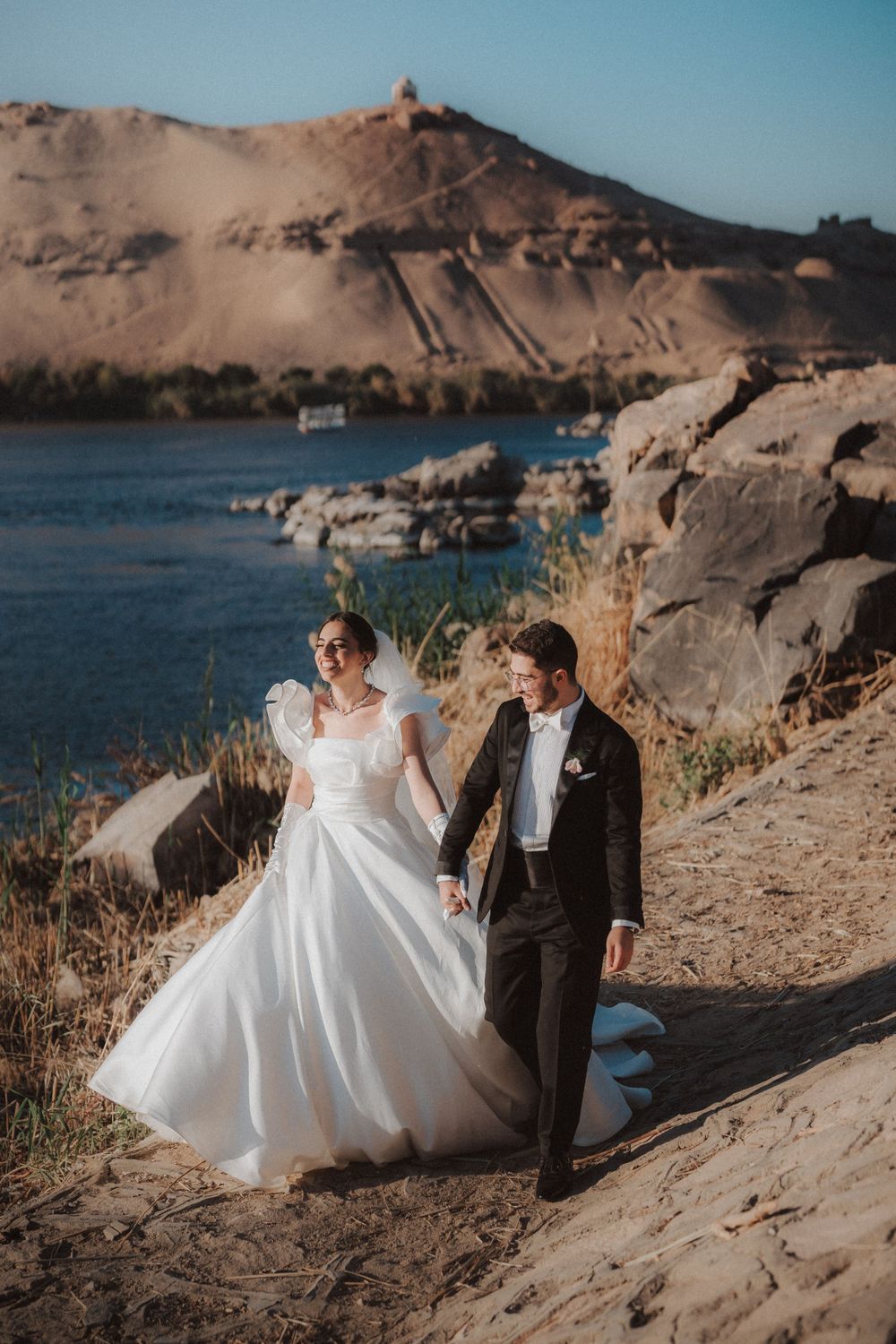 A couple in wedding attire walks along a scenic riverside path with desert mountains in the background.