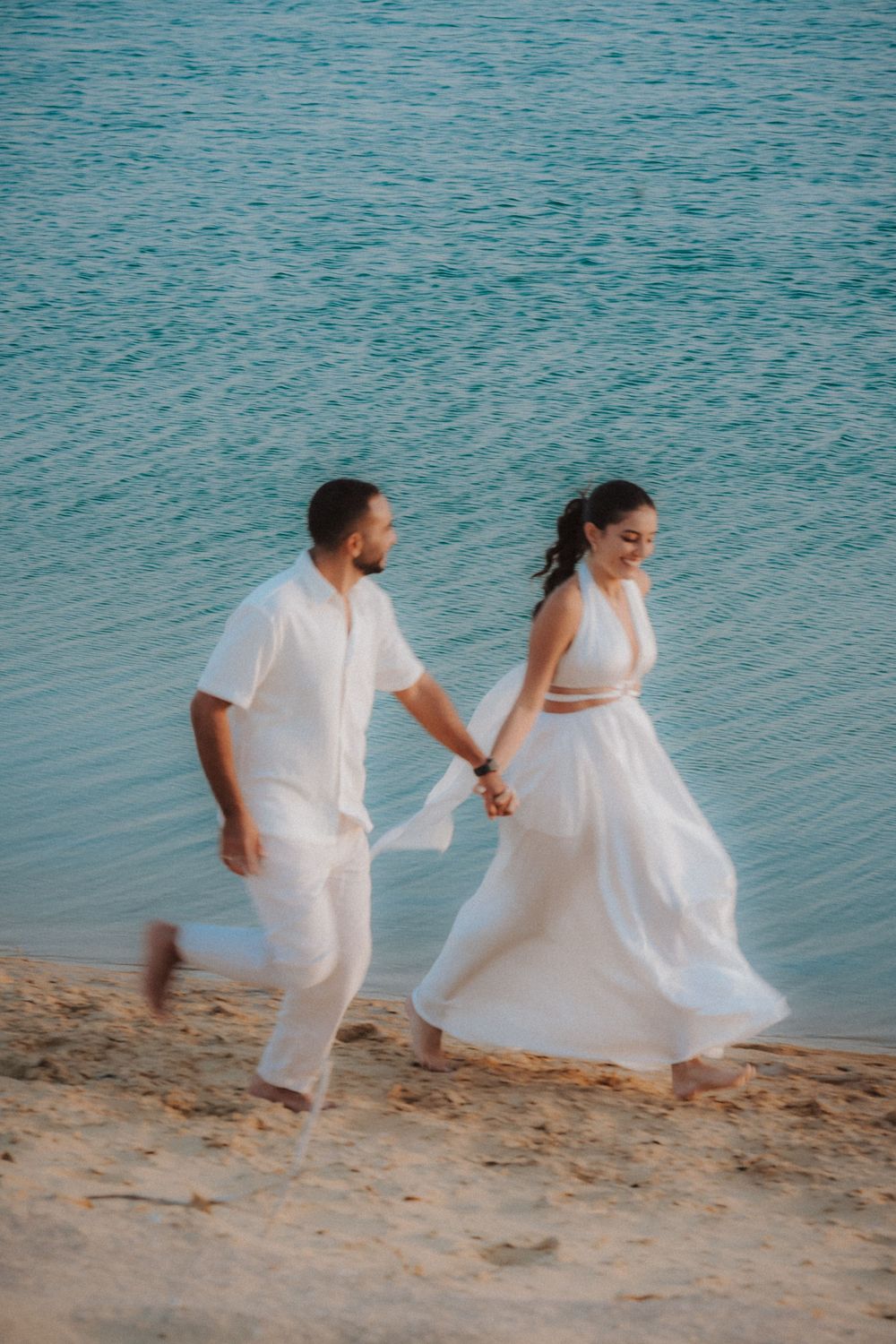Joyful couple in white attire running along turquoise water beach at sunset.