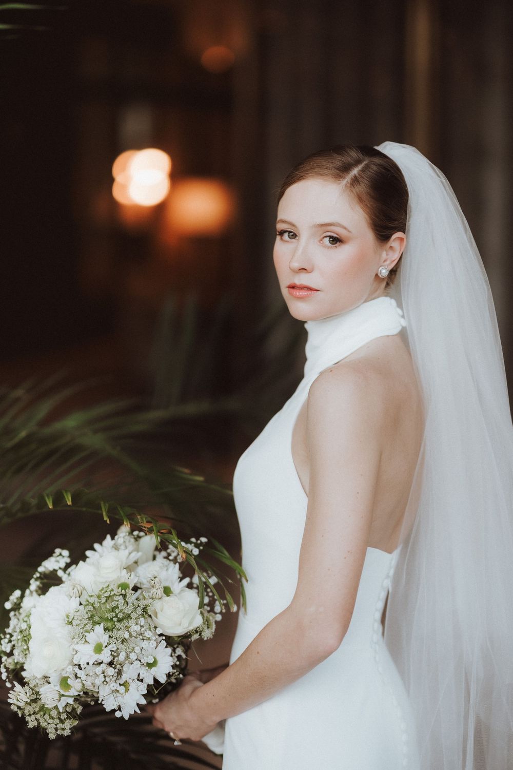 A bride in a white wedding dress and veil poses for romantic bridal portraits in a dimly lit indoor setting.