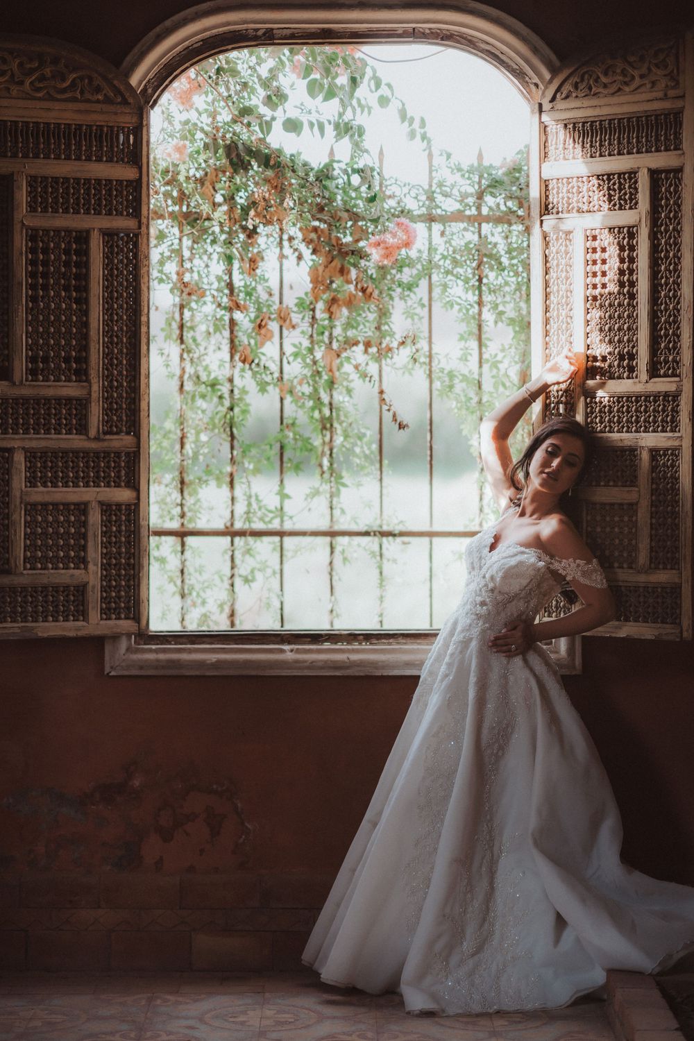A flowing white wedding gown glows in soft light by an ornate window with climbing vines and latticework details.