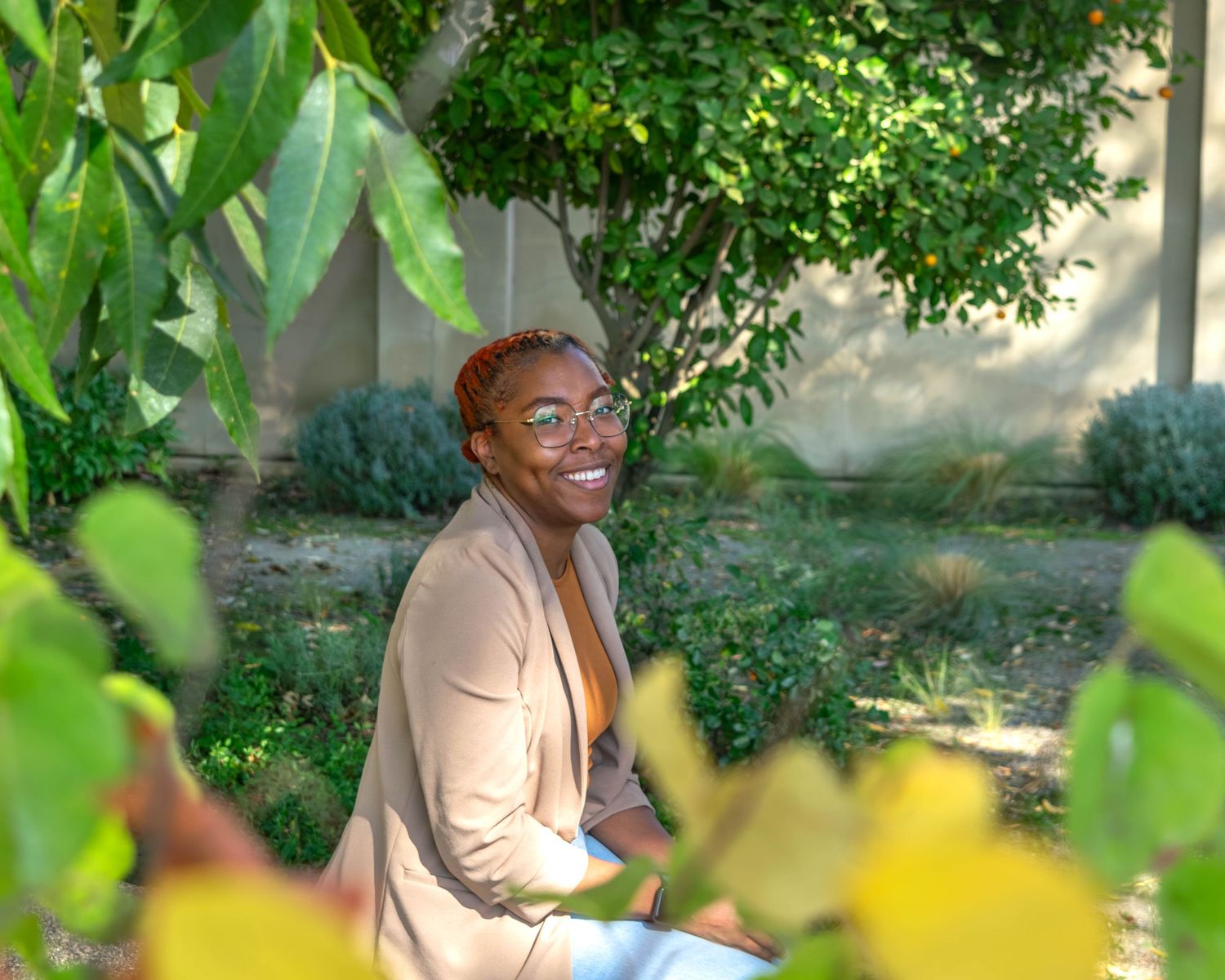 Person in beige blazer sitting outdoors surrounded by lush green foliage.