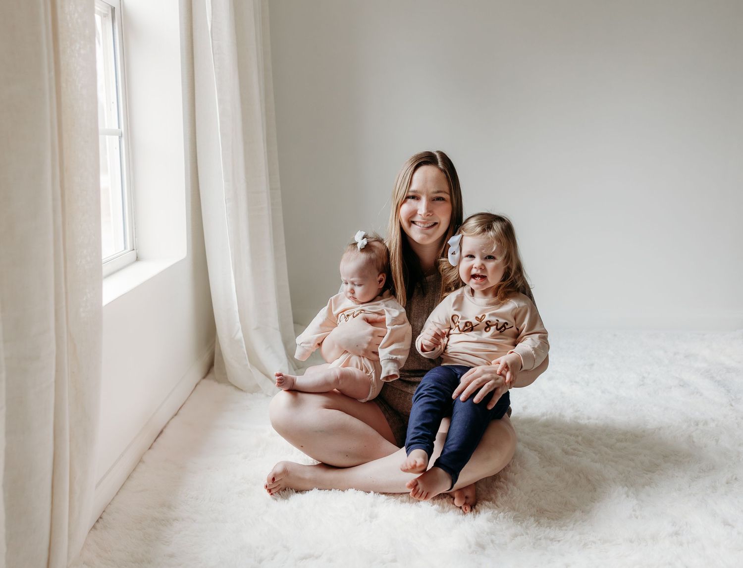 Smiling mother sits cross-legged on a fluffy rug holding two young daughters in a bright airy studio.