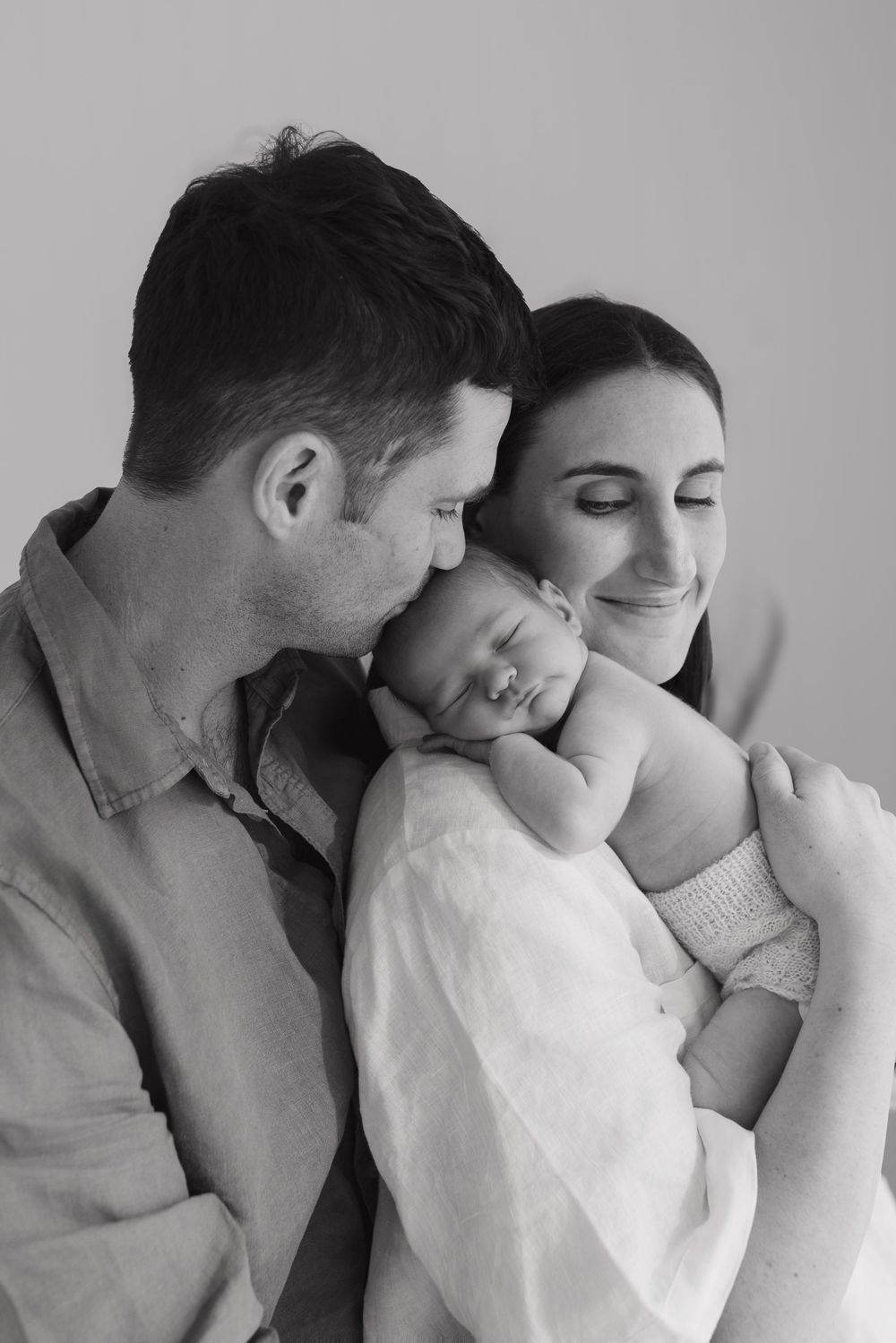 A tender black and white portrait of a couple cradling their sleeping newborn baby in an intimate family moment.