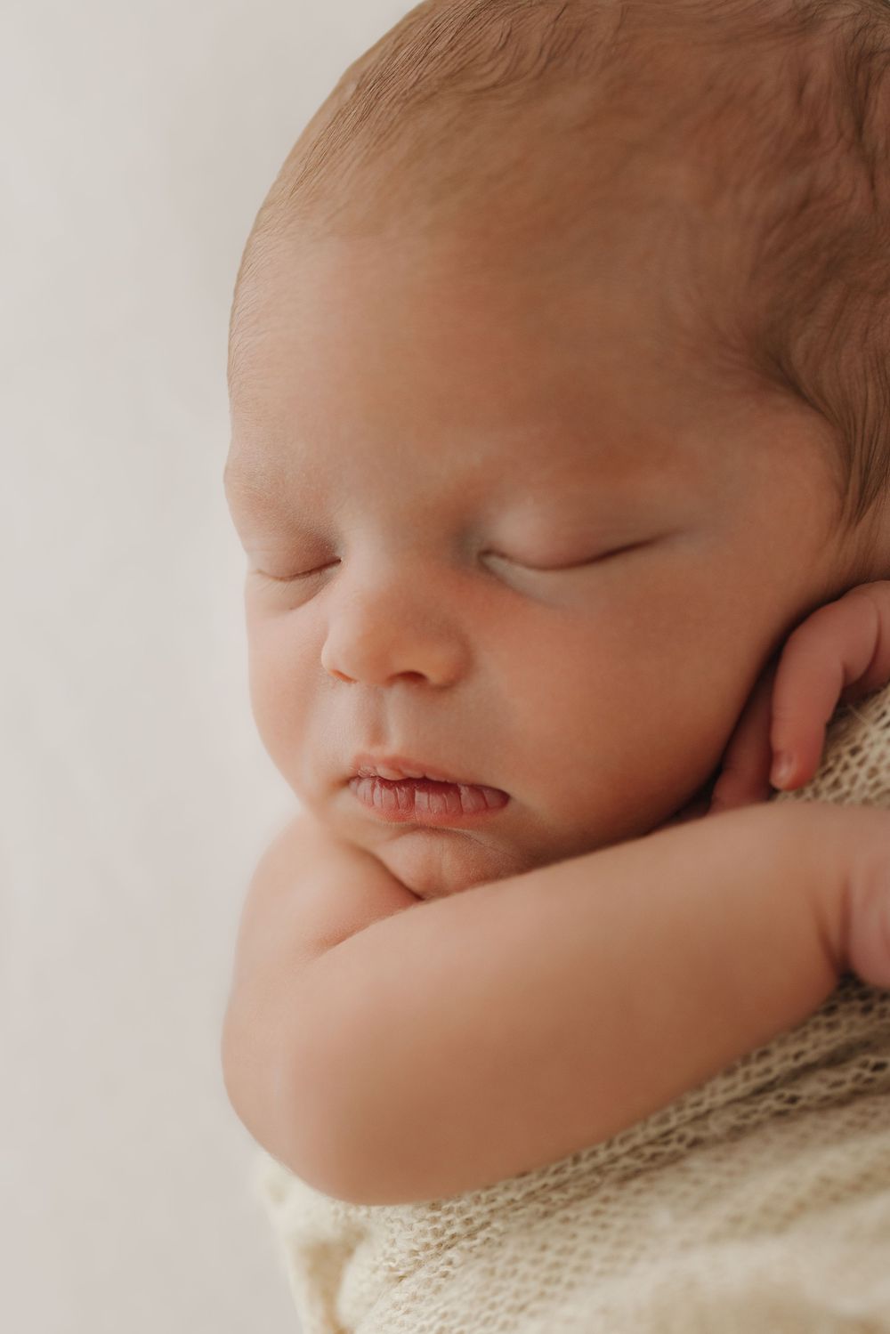 Close-up profile view of a peaceful sleeping newborn wrapped in cream-colored fabric.