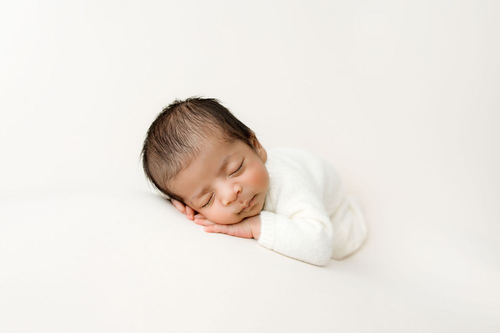 A tiny newborn baby boy dresses in a white romper lying on his tummy with head on hands. A simple all white/creamy baby portrait