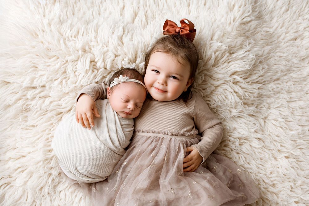 The cutest sweet two sisters, toddler and a wrapped newborn baby, both lying down and big sister smiling at the camera