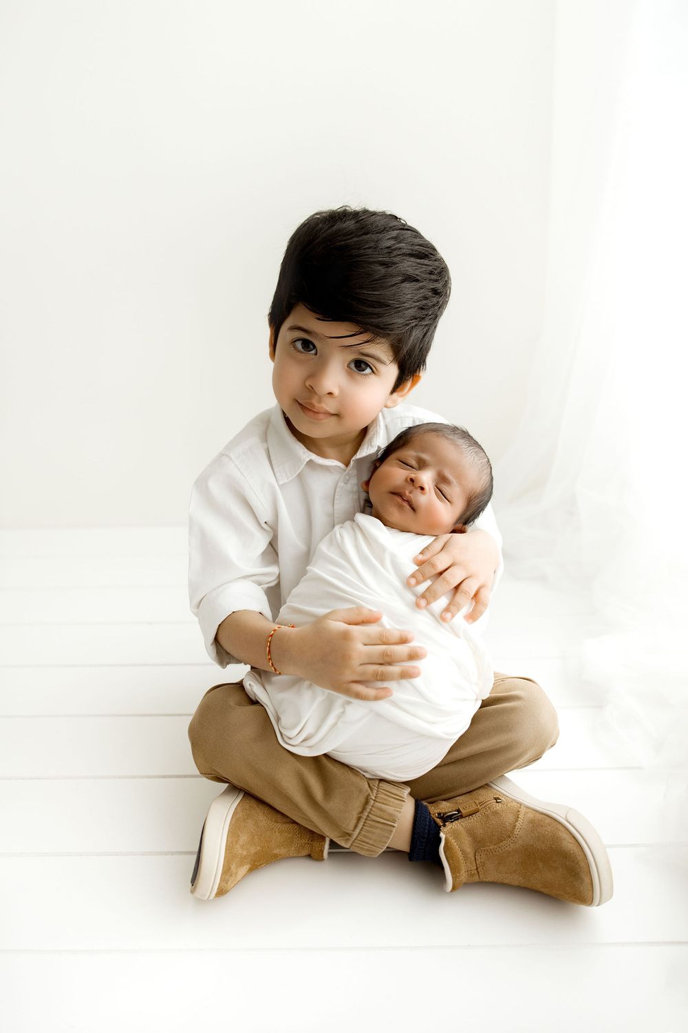 Proud three years old big brother holding his newborn baby brother in his lap, studio session by the window
