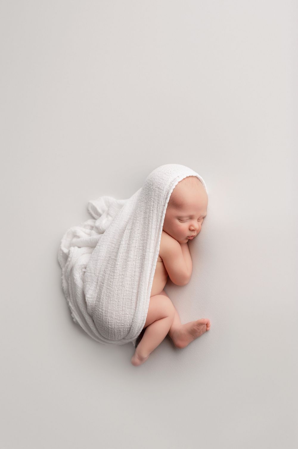 Peaceful newborn baby sleeping wrapped in a soft white muslin swaddle blanket on a light gray background.