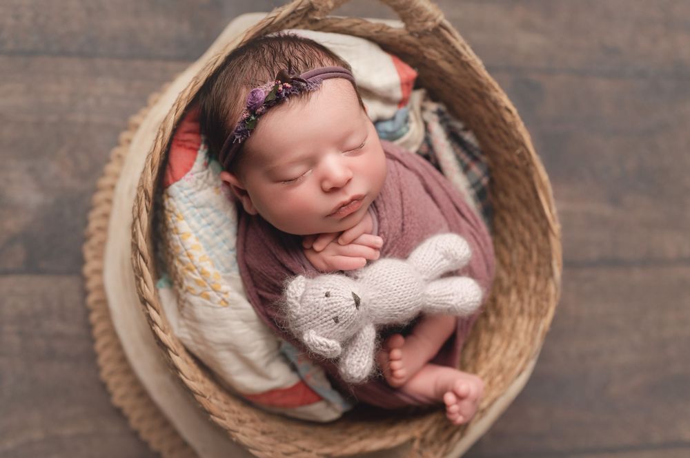 Newborn baby girl sleeping in a wicker basket wrapped in purple with a knit bunny stuffed animal.