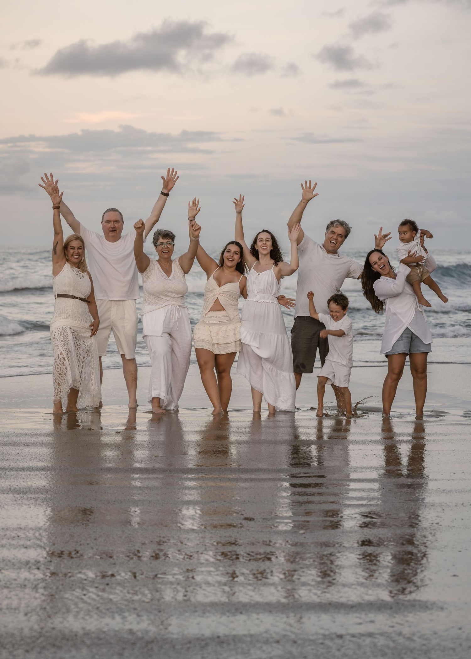 A group wearing white outfits poses playfully together at the beach during sunset with waves in the background.