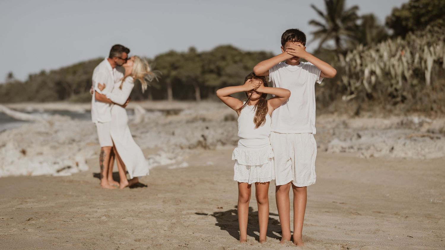 Two couples in white outfits embrace while standing on a sandy beach at sunset.