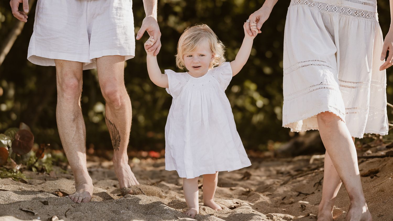 A toddler in white holding hands with parents while walking on sandy beach.