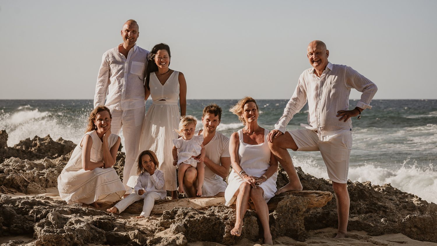 A family portrait on rocky beach terrain dressed in coordinated white outfits at sunset.
