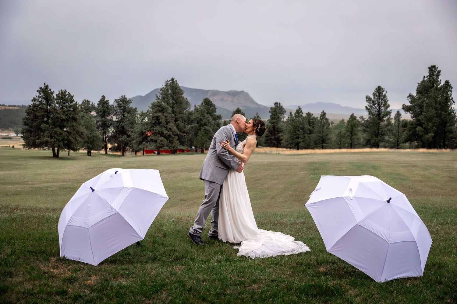 Newlyweds share romantic kiss by serene lake with mountains in background.