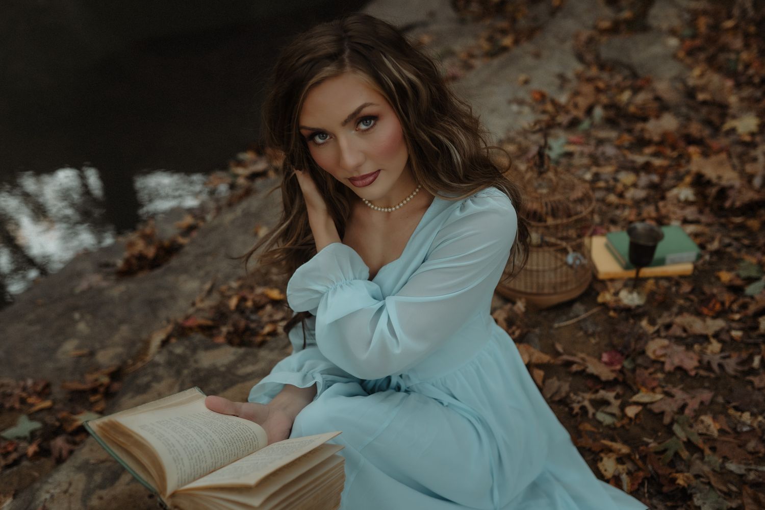 Someone in a light blue dress sits on autumn leaves reading a book in moody outdoor lighting.