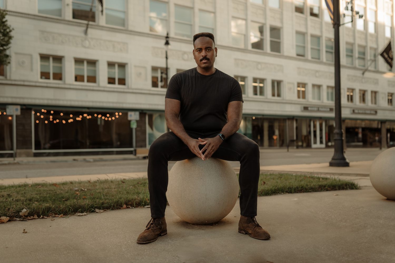 Person in black clothing sits on concrete steps in front of building with string lights visible in background.