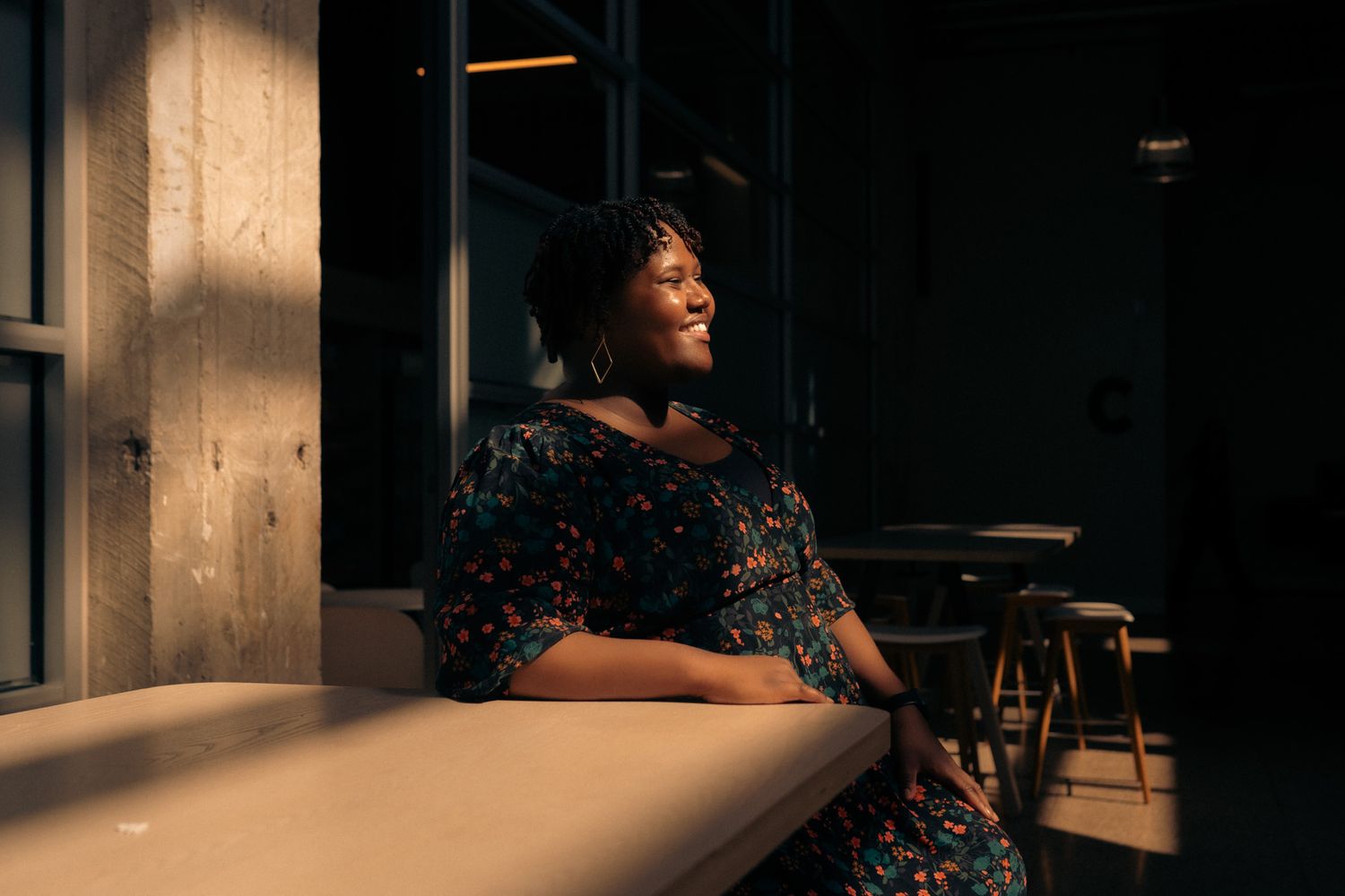 A person in a floral dress sits at a table in warm lighting near a window.
