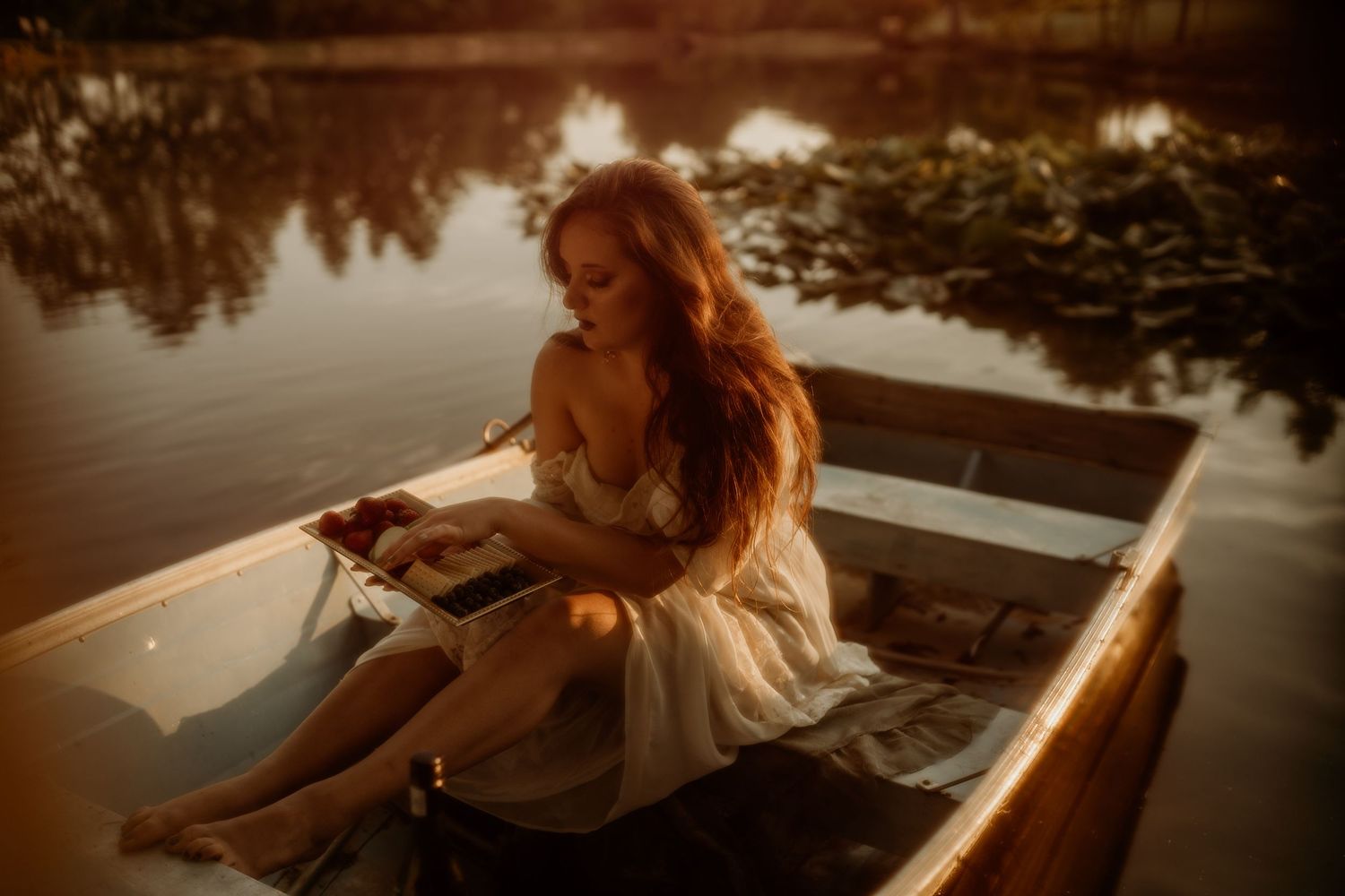 A person in a white dress reads peacefully in a wooden boat on calm waters at sunset.