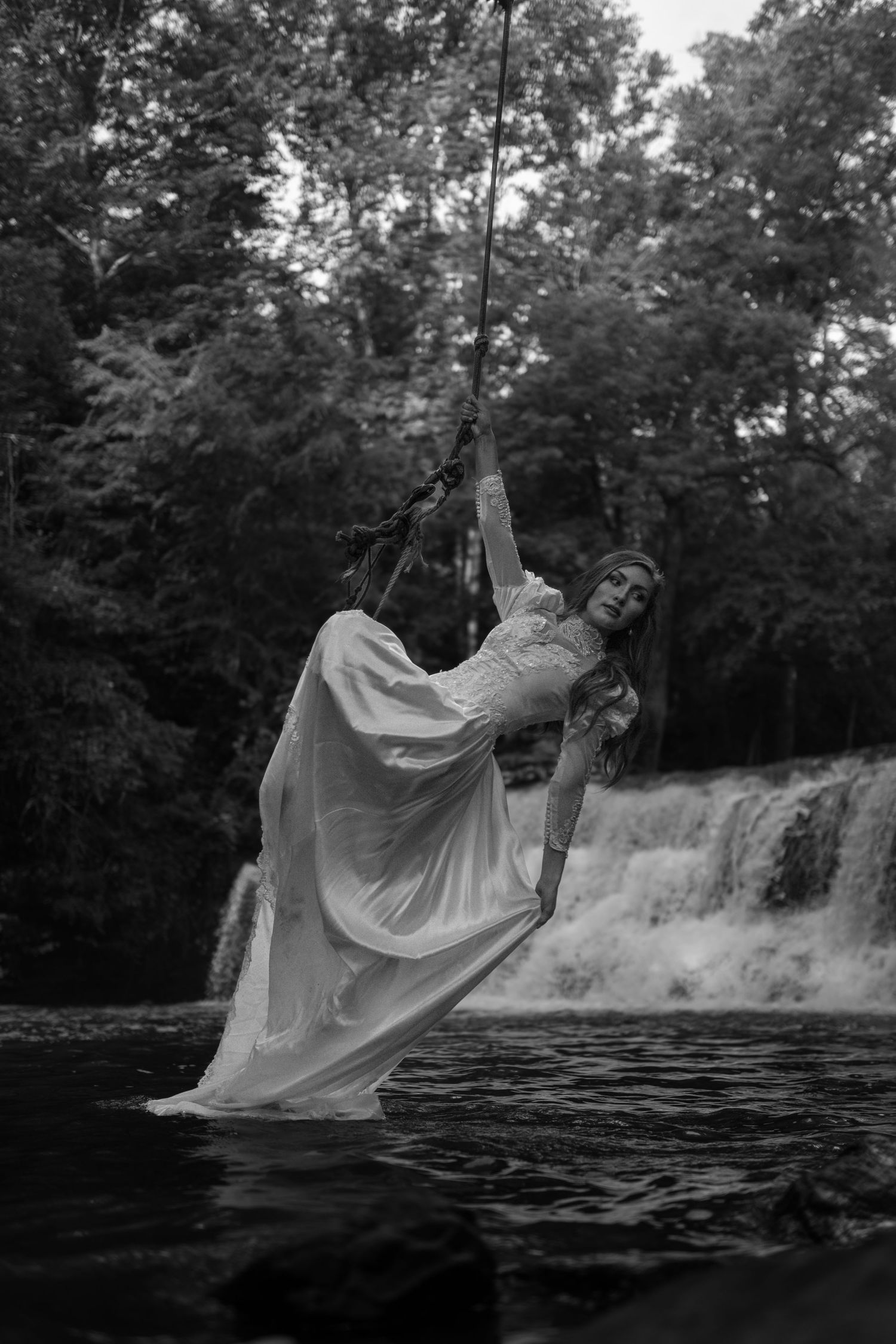 Black and white photo of a woman in a white gown swinging on a rope over a river with a waterfall backdrop.