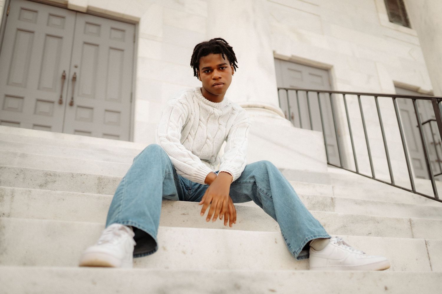Person in light blue jeans and white t-shirt sits casually on white marble steps in an outdoor setting.