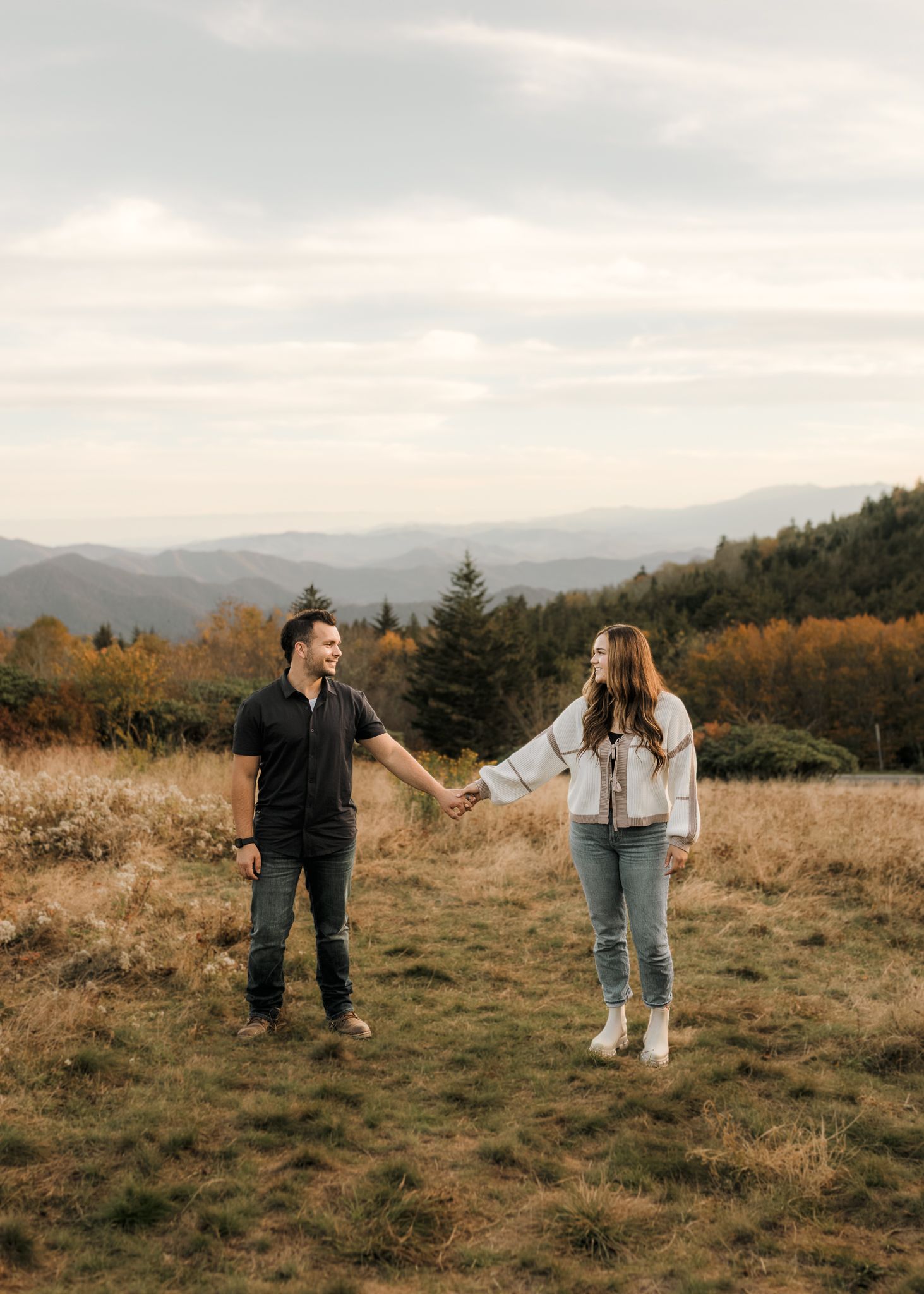 Two people hold hands while standing in a grassy field with mountains and autumn trees in the background at sunset.